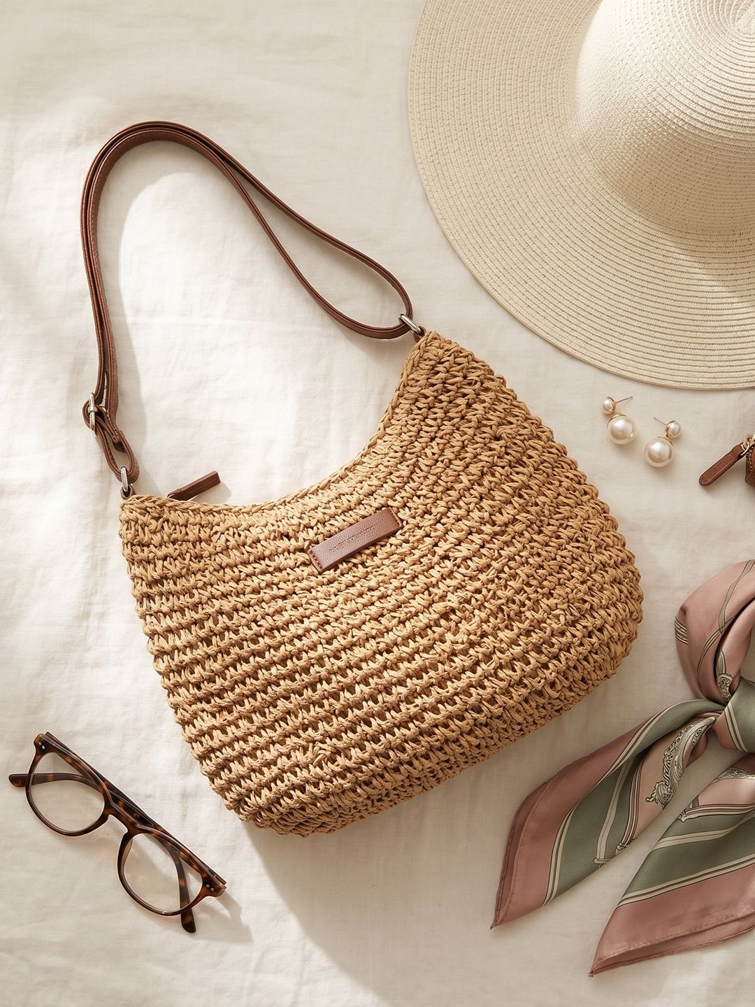 Bolso bandolera hobo trenzado beige con asa larga de cuero marrón y detalle rectangular.