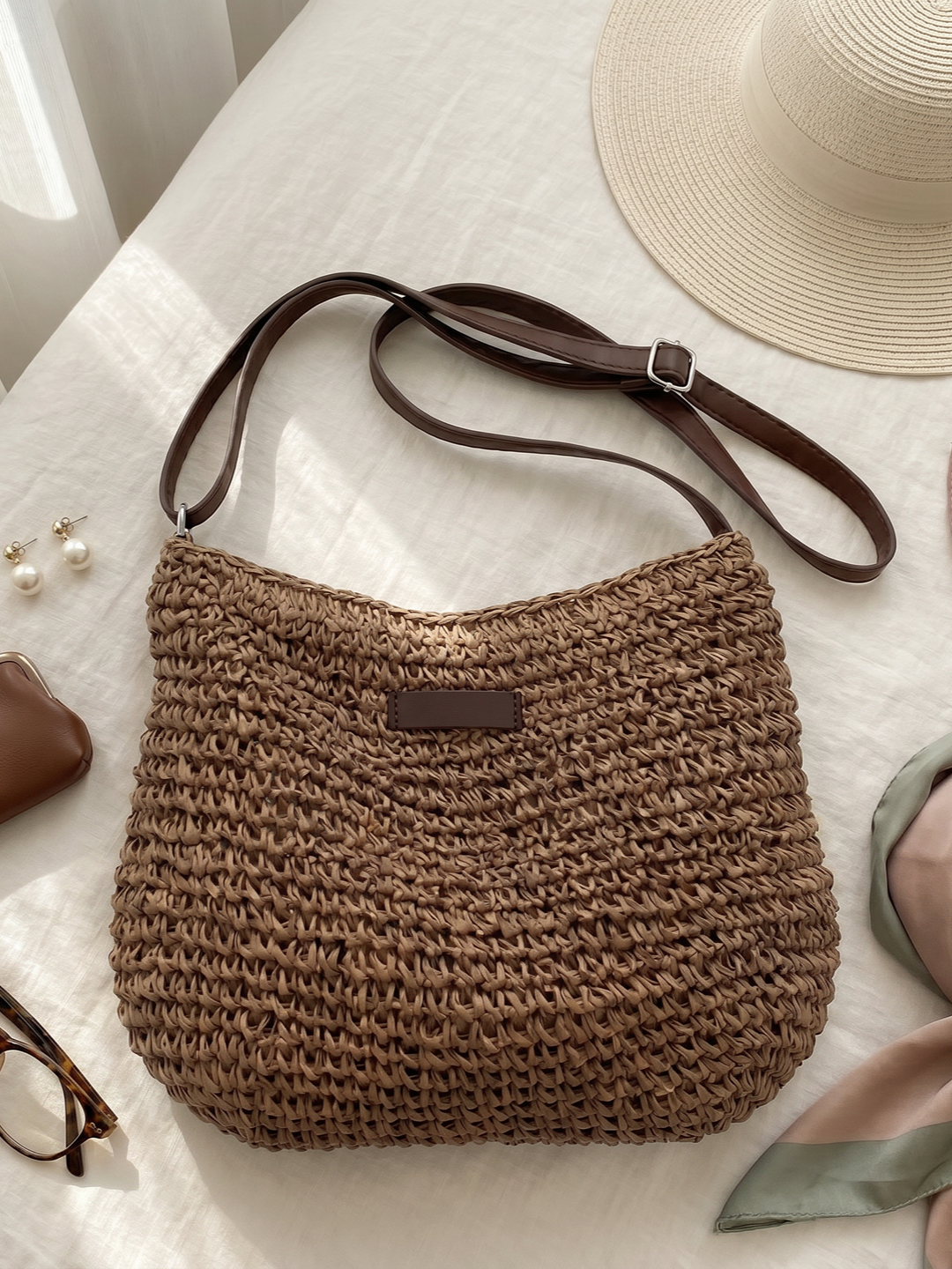 Bolso hobo de rafia trenzada beige con asa de cuero marrón y cierre frontal rectangular.